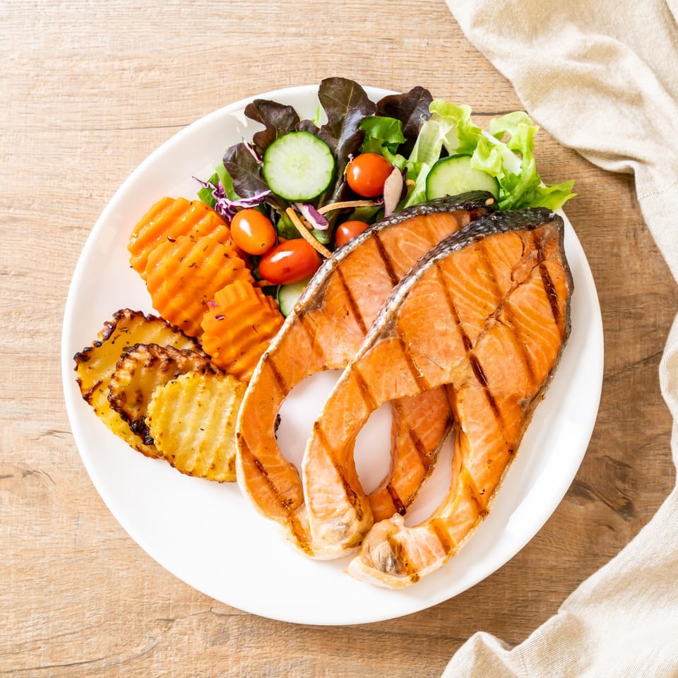Grilled salmon steaks with a side of fresh salad, roasted carrots, and potatoes on a white plate.