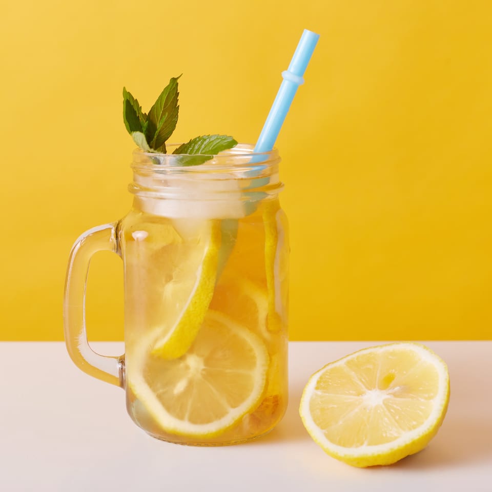 Lemonade in a mason jar with lemon slices, mint, and a blue straw, next to a lemon half.