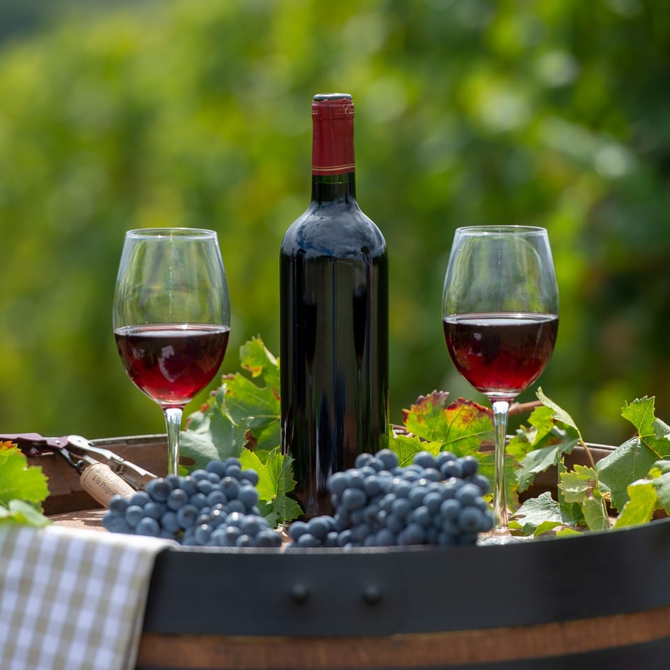 Red wine bottle and two glasses with grapes and corkscrew on a barrel, vineyard in background.