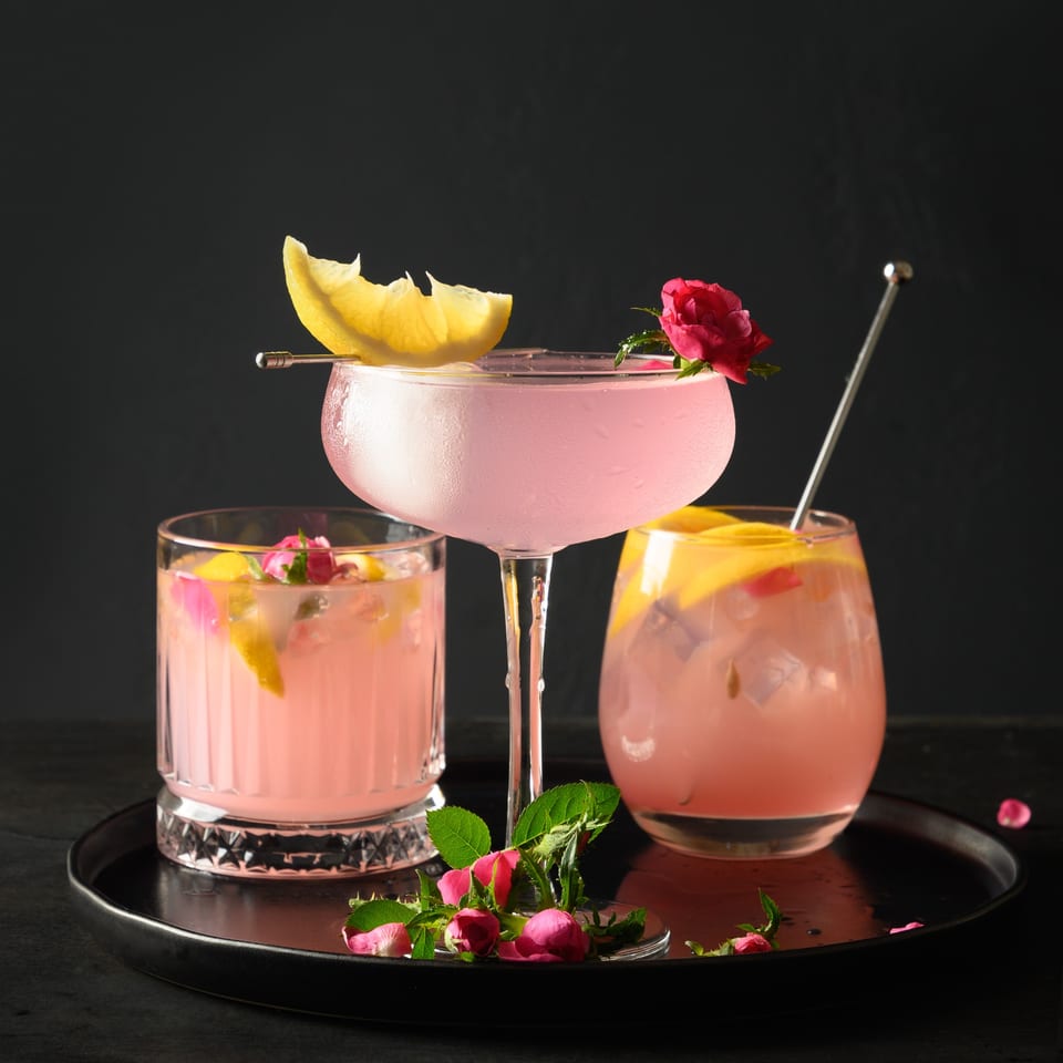 Three pink cocktails garnished with lemon slices and rosebuds on a black tray.