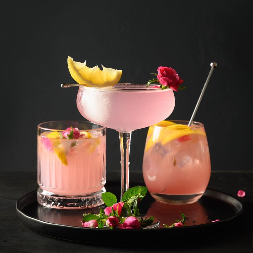 Three pink cocktails garnished with lemon slices and rosebuds on a black tray.